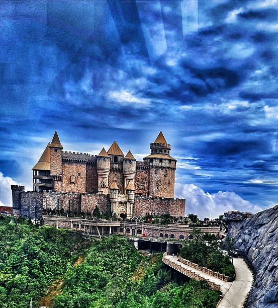 Danang, Vietnam on 12 May 2025. Ba Na Hills. Stone castle with towers stands atop a green hilltop under a dramatic cloudy blue sky, creating a fantasy atmosphere with winding roads, ideal for travel and historical themed materials.