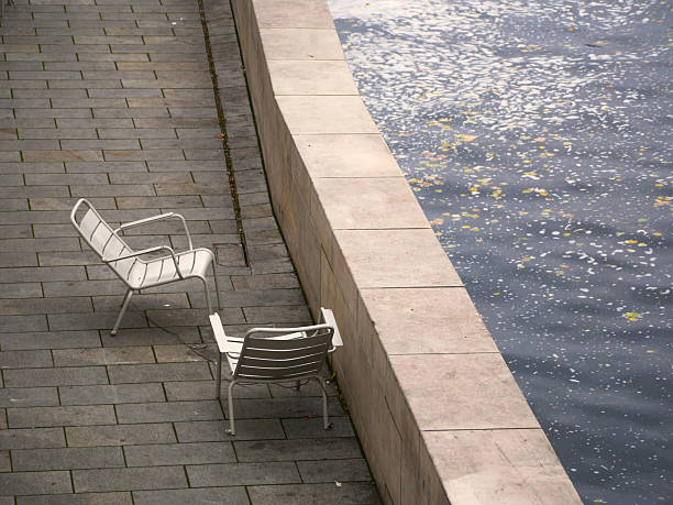 two empty metallic foldable chairs standing on the pavement close to the shore of a river in a city