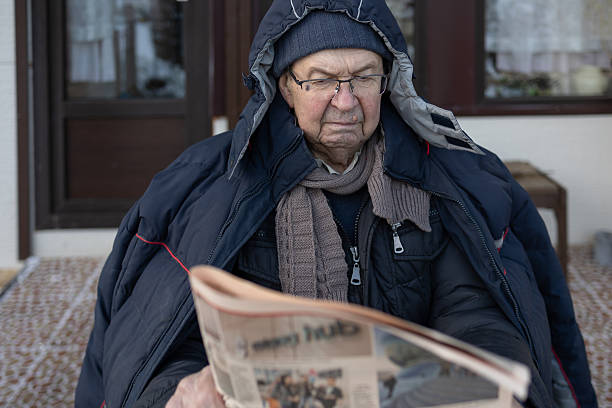 Elderly man in winter clothing reading newspaper at outdoor table with cup and bowl. Calm domestic setting on natural light.