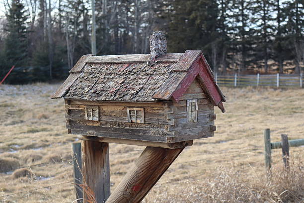A mailbox fastened on a post beside a road on a rural area. The mailbox is built like a small log cabin and looks very rustic. The paint has long since worn of.