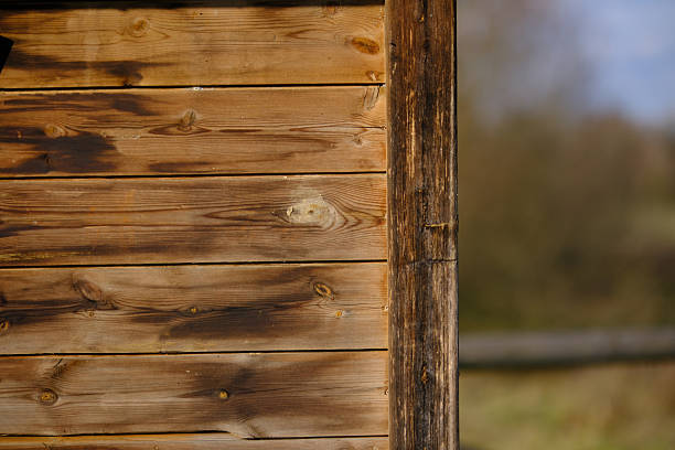 Close-up detail of a rustic wooden wall with visible grain, knots, and natural aging marks. The vertical post contrasts with horizontal planks, while a softly blurred outdoor background adds depth. Ideal for concepts such as craftsmanship, sustainability, rural architecture, natural materials, and minimal design.
