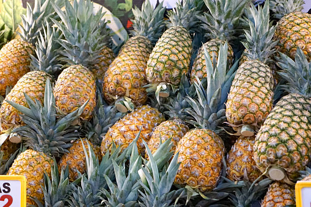 A dense pile of fresh pineapples with vivid yellow-green skin and spiky leaves, stacked in various angles at a bustling market or grocery store.