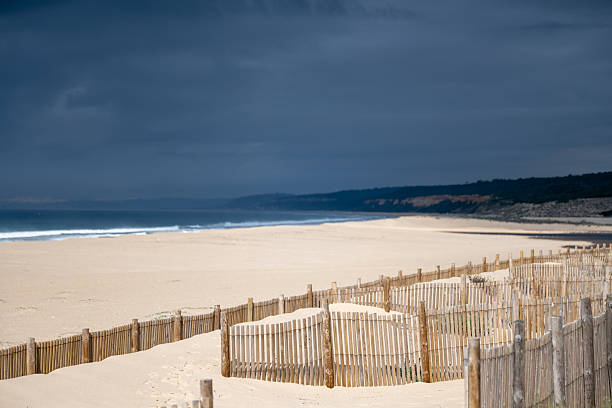 Wooden sand fences run across coastal dunes beside a wide Atlantic beach under dark clouds at Lagoa de Albufeira Sesimbra Portugal on December 25 2025.
