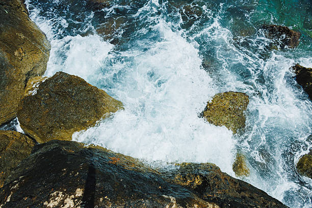 Powerful waves break against the jagged rocks near the coast, creating a beautiful display of foam and spray in the bright midday sun. The water is clear, showcasing vibrant shades of blue.