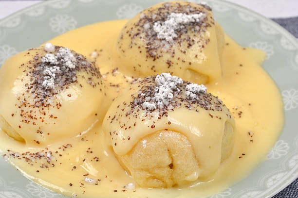 A close up shot of three sweet yeast dumplings topped with thick vanilla sauce poppy seeds and powdered sugar on a decorative plate