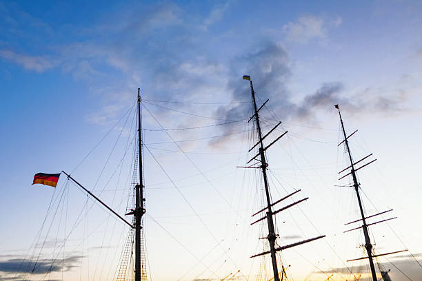 Three tall ship masts rise against sunset sky, rigging silhouetted in warm orange and blue hues. German flag flutters on leftmost mast, adding a touch of national identity to the nautical scene