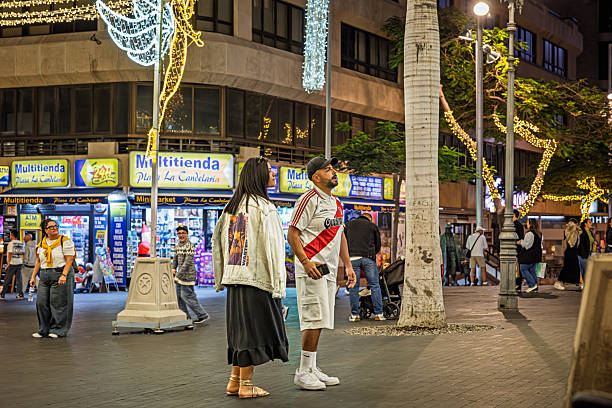 Santa Cruz, Tenerife, Spain, Europe - December 8th 2025: A young couple looking in amazement at the extensive Christmas decorations hanging between tall palm trees on the central square of an old Spanish town. The scene captures the contrast between festive holiday lights and the warm southern atmosphere, highlighting the charm of seasonal celebrations in a historic subtropical setting.