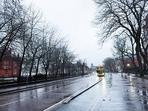Manchester, UK - December 06, 2025: Damp city street in Manchester with a bright bus, bare trees, and moody skies.
