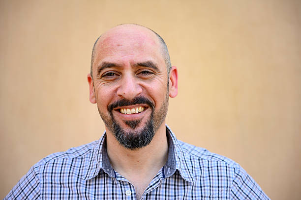 Confident mid adult man with a bald head and goatee smiling widely while standing against a neutral background, expressing happiness and approachability