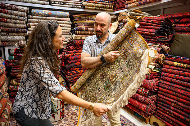 Customer and merchant inspecting a traditional handmade carpet, surrounded by stacks of colorful textiles and authentic rugs, highlighting craftsmanship and local shopping experience
