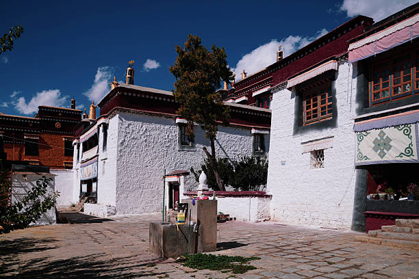 A wide-angle view of the historic Sera Monastery in Lhasa, Tibet, showcasing its iconic white-washed stone walls, red clay decorative friezes, and traditional Tibetan windows. The sacred courtyard is bathed in bright Himalayan sunlight under a deep blue sky with scattered white clouds. This image captures the serene and majestic atmosphere of one of the \