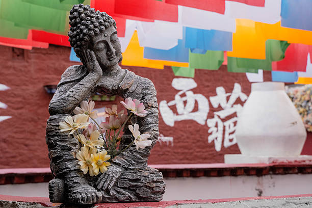 A contemplative stone Buddha statue rests peacefully, adorned with delicate flowers in its hollow center. The background features traditional colorful Tibetan prayer flags and a red monastery wall with Chinese characters for \