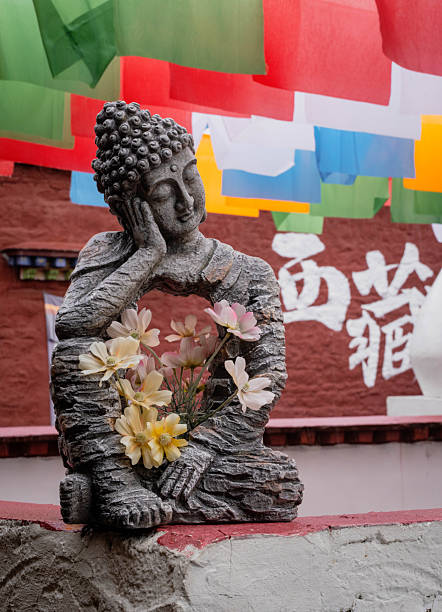 A contemplative stone Buddha statue rests peacefully, adorned with delicate flowers in its hollow center. The background features traditional colorful Tibetan prayer flags and a red monastery wall with Chinese characters for \