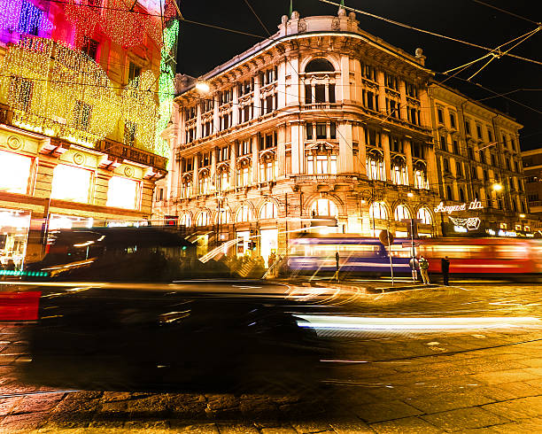 A dynamic night scene in the city center of Milan, Italy, featuring a white tram passing through the street. Festive holiday light decorations hang above the road, framed by historic buildings and brightly lit shop windows. Pedestrians are seen walking along the sidewalks, capturing the busy urban atmosphere of the winter season. The image highlights the blend of public transport and iconic Gothic architecture in the Lombardy capital.