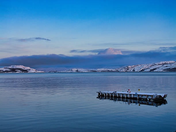 A quiet winter view of Kirkenes Bay in Kirkenes, Norway, with a small dock on calm water, snowy hills, and soft blue-hour skies.