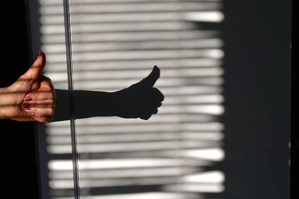A play of shadows. The shadow of a hand, with a raised thumb, and window blinds is projected onto the wall.