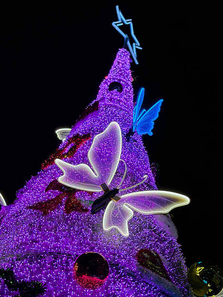 Stock photo shows an artificial pink spruce Christmas tree with realistic fake needles, adorned with butterfly ornaments and colorful baubles, set outdoors at night. The glowing, whimsical scene creates a magical and festive holiday atmosphere