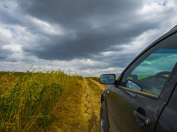 Car navigates a dirt road surrounded by tall grass fields under a cloudy sky. The scene evokes a sense of adventure and solitude, creating a dramatic atmosphere.