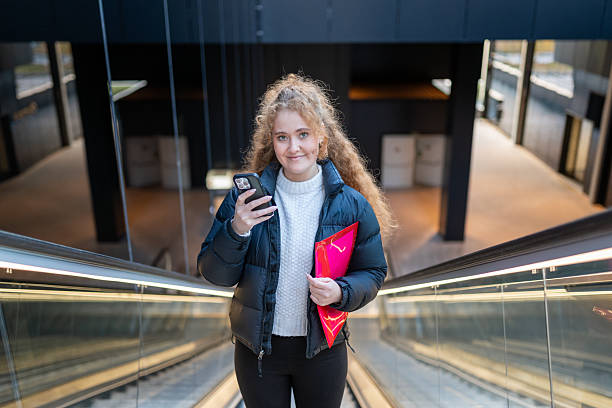 istock Young woman commuting on escalator, smiling and holding smartphone with red folder, navigating urban environment 2254259177