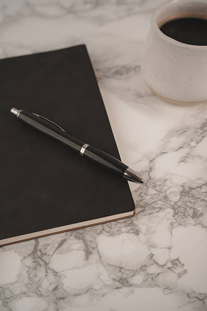 istock A black notebook lies open with a pen resting on top, accompanied by a white coffee cup filled with dark coffee. This setup invites creativity and reflection 2254269381