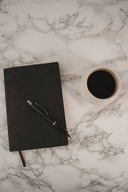 istock A black notebook lies open beside a sleek pen on a smooth marble surface. Next to it sits a white cup filled with dark coffee, inviting moments of inspiration and creativity 2254269446