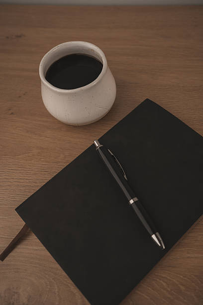 A black notebook and pen rest on a smooth marble table beside a white coffee cup filled with dark coffee. This scene captures a peaceful moment for creativity