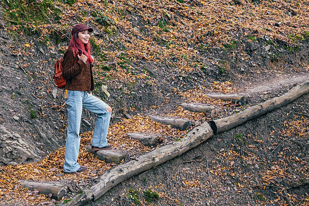 Young woman with backpack climbing rustic log steps on a vibrant autumn forest trail, smiling amid fallen leaves and foliage