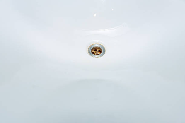 A circular metal sink drain sits centered on white porcelain with slight rust around the grate, indoors, using high key lighting and negative space for a minimalist look.