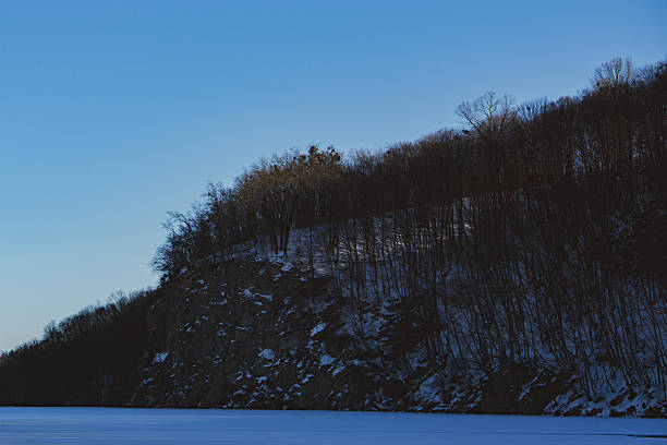 A frozen winter river stretches past rocky cliffs covered with trees, all blanketed in white snow. The bright blue sky and sunshine create a crisp, peaceful winter atmosphere.