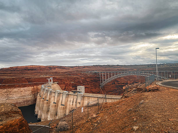 A wide view of the Glen Canyon Dam and the adjacent bridge, set against the backdrop of arid red rock formations and a moody sky