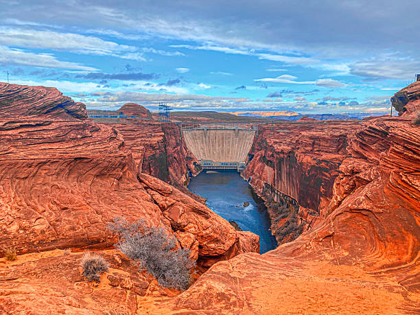 A wide view of the Glen Canyon Dam and the Colorado River surrounded by vibrant red sandstone cliffs and a dramatic cloudy sky
