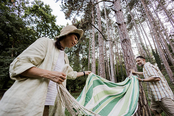 Couple setting up a hammock while preparing for a camping trip in nature. Man and woman enjoying outdoor adventure, teamwork, and relaxation before settling into their campsite. Concept of camping lifestyle, travel, leisure time, and connection with nature.