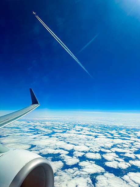 Above the soft white clouds, a jet plane leaves a trail in the bright blue sky while the airplane wing stands out against the vast horizon during daytime travel.