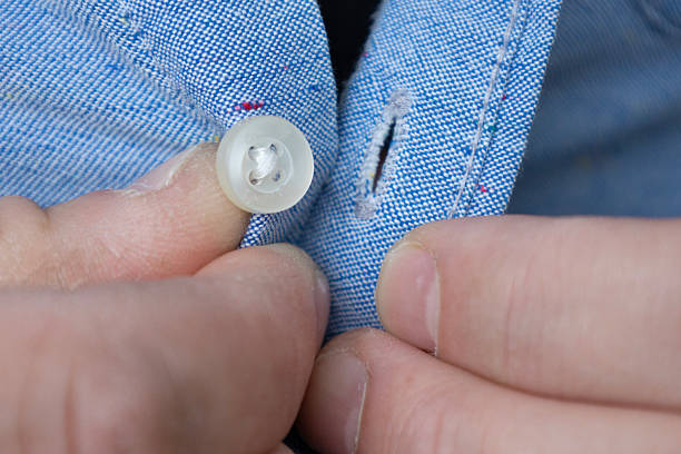 Close-up of hands fastening a blue cotton shirt button, showing fabric texture, stitching and casual clothing detail in everyday dressing concept.