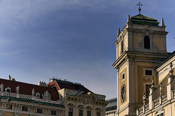 An architectural detail shot focusing on the yellow stucco facade and green roof of a historic baroque tower and adjacent buildings, featuring statues along the rooftop, set against a deep blue, clear sky on a sunlit day, likely in Vienna, Austria.