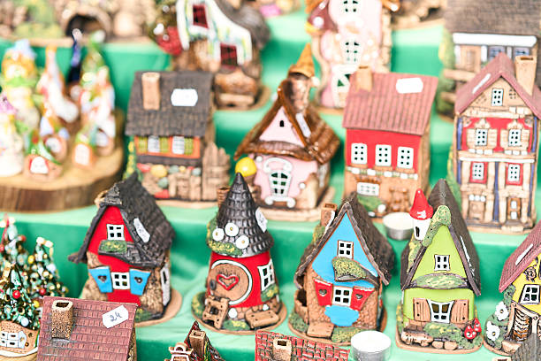 A close-up shot captures a vibrant collection of handmade ceramic miniature houses, painted in bright reds, blues, and greens, displayed closely together on a craft market stall.