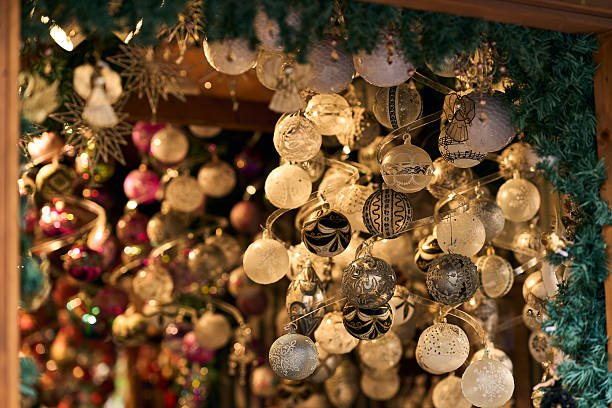 A festive tangle of intricately decorated white, silver, and patterned glass Christmas ornaments hanging from a market stall at a holiday fair, featuring bright colorful bokeh lights in the warm background.
