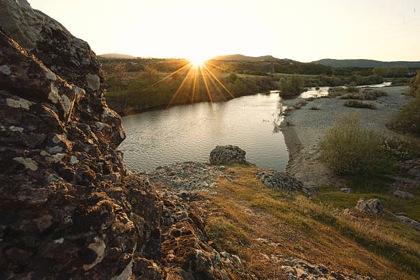 Scenic river flowing through a tranquil valley at sunset, with striking rocks in the foreground and gentle hills rising in the background. Sun reflections in the water