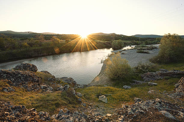 Scenic river flowing through a tranquil valley at sunset, with striking rocks in the foreground and gentle hills rising in the background. Sun reflections in the water