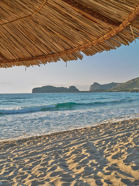 Photo from the island of Crete (Greece): A tranquil Mediterranean beach scene shows a woven straw thatched awning framing the foreground, with clear turquoise water, gentle waves, and warm golden sand. In the distance, rugged islands rise against a soft sky, conveying calm, vacation vibes, and timeless coastal beauty in Greece.