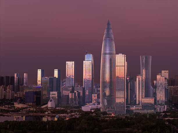 The modern skyline of Shenzhen, featuring the iconic Ping An Finance Center rising above the central business district. Contemporary skyscrapers glow in warm evening light under a purple dawn sky, reflecting the rapid urban development and economic growth of southern China.