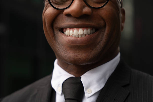 A man wearing a suit and tie stands outdoors with a big smile. The background shows a city scene during daylight. He appears happy and confident.