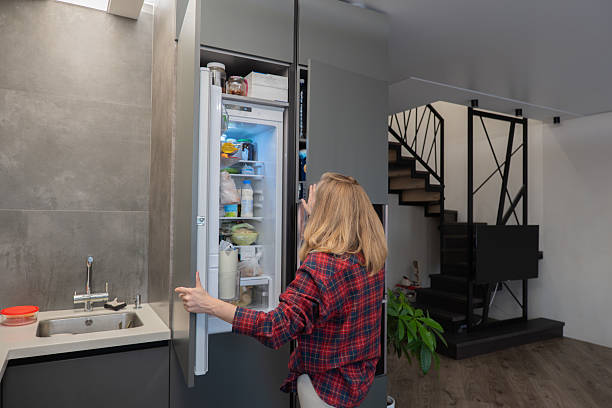istock Woman opening refrigerator in modern kitchen, freelance designer grabbing quick snack between remote calls, wearing plaid shirt, blonde hair, openplan living area with staircase and media setup, 2254321617