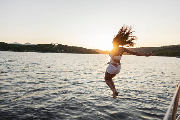Young woman leaps from a boat into a lake at golden hour, backlit silhouette capturing summer freedom, joy and spontaneous vacation adventure on calm water