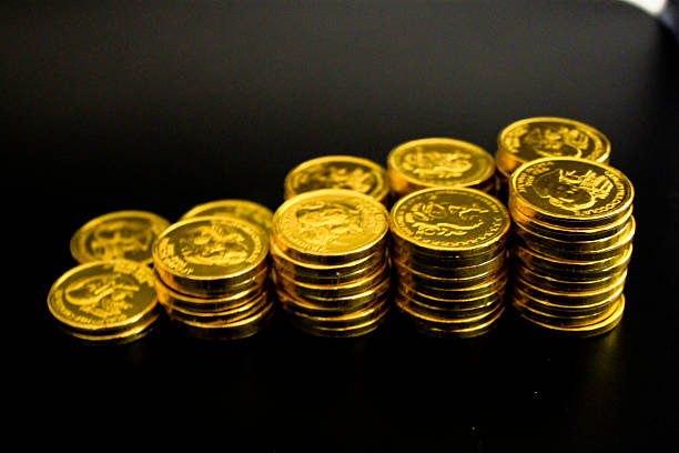 A close-up view of shiny, foil-wrapped chocolates designed to resemble gold coins. The coins are stacked in small piles and scattered across a smooth surface, their metallic golden wrappers glistening under soft light. Each wrapper features embossed details mimicking real currency, adding a realistic touch.