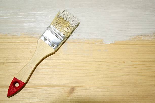 istock Wooden paintbrush with a red tip rests on a light wood surface that is half-covered in a white paint coat, illustrating the process of home improvement and DIY renovation 2254327622