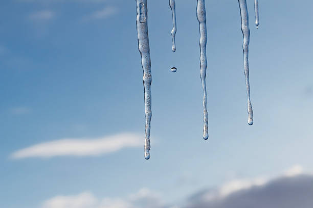 Transparent icicles hanging from roof edge with single water drop falling, soft blue sky and clouds in background, concept of thaw, changing seasons and winter weather.