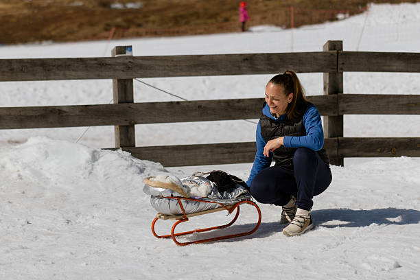 Woman dressed in warm winter clothing checking a sled with her baby before sledding, surrounded by snow and wooden fence at a winter ski destination