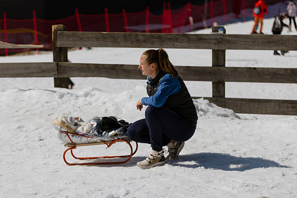 Caring mother kneeling in the snow while securing her baby on a sled at a winter mountain resort