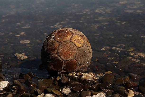 An old, worn-out soccer ball rests partially submerged among barnacle-covered rocks in shallow water, evoking themes of abandonment, memory, and the passage of time.